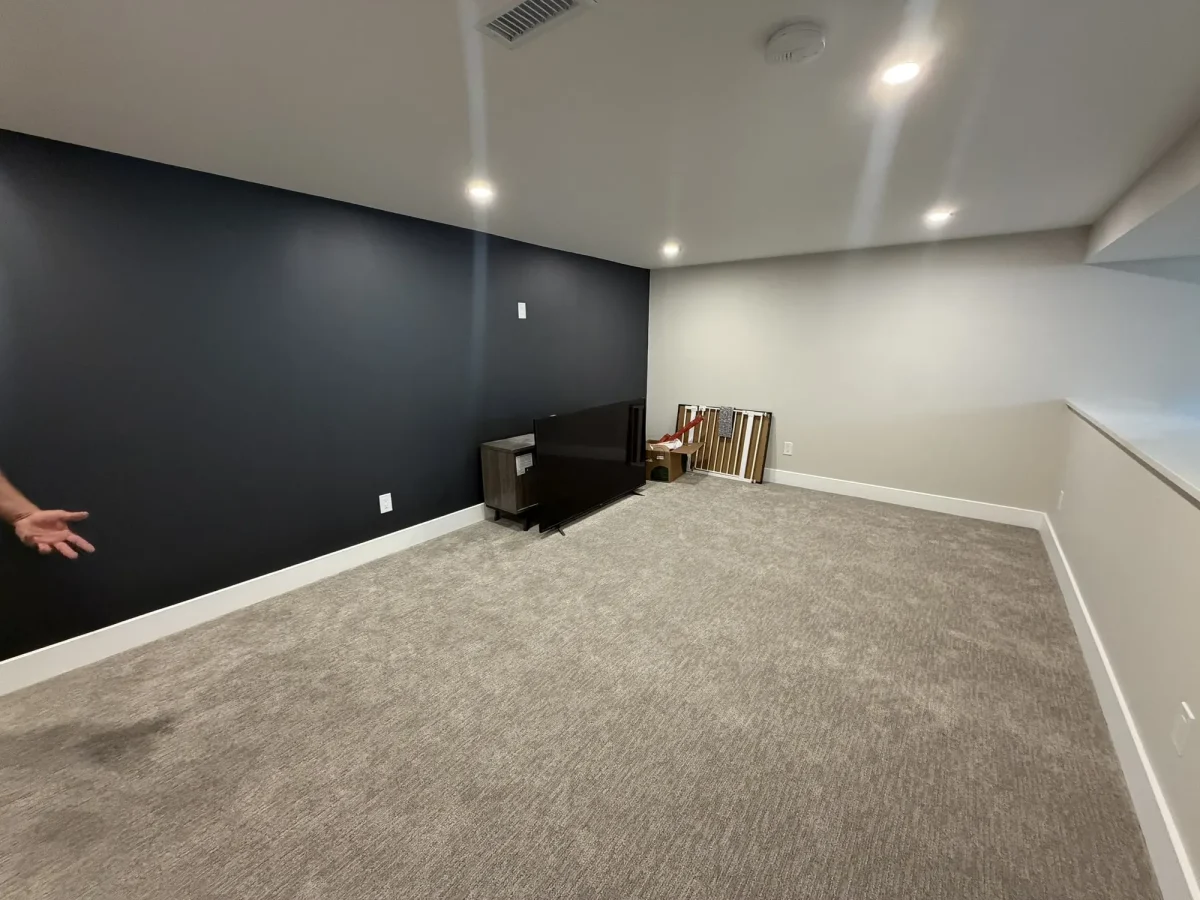 Finished basement room with a charcoal feature wall, fresh carpet and recessed ceiling lights.
