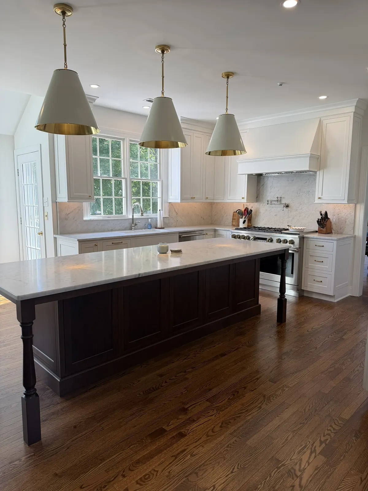 White kitchen with a dark-stained island under three brass cone pendants, framed by a window over the sink.