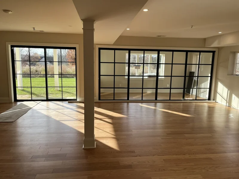 Great room in a newly built home with black steel-framed window walls along two sides, a white column, and wide oak floorboards catching afternoon light.