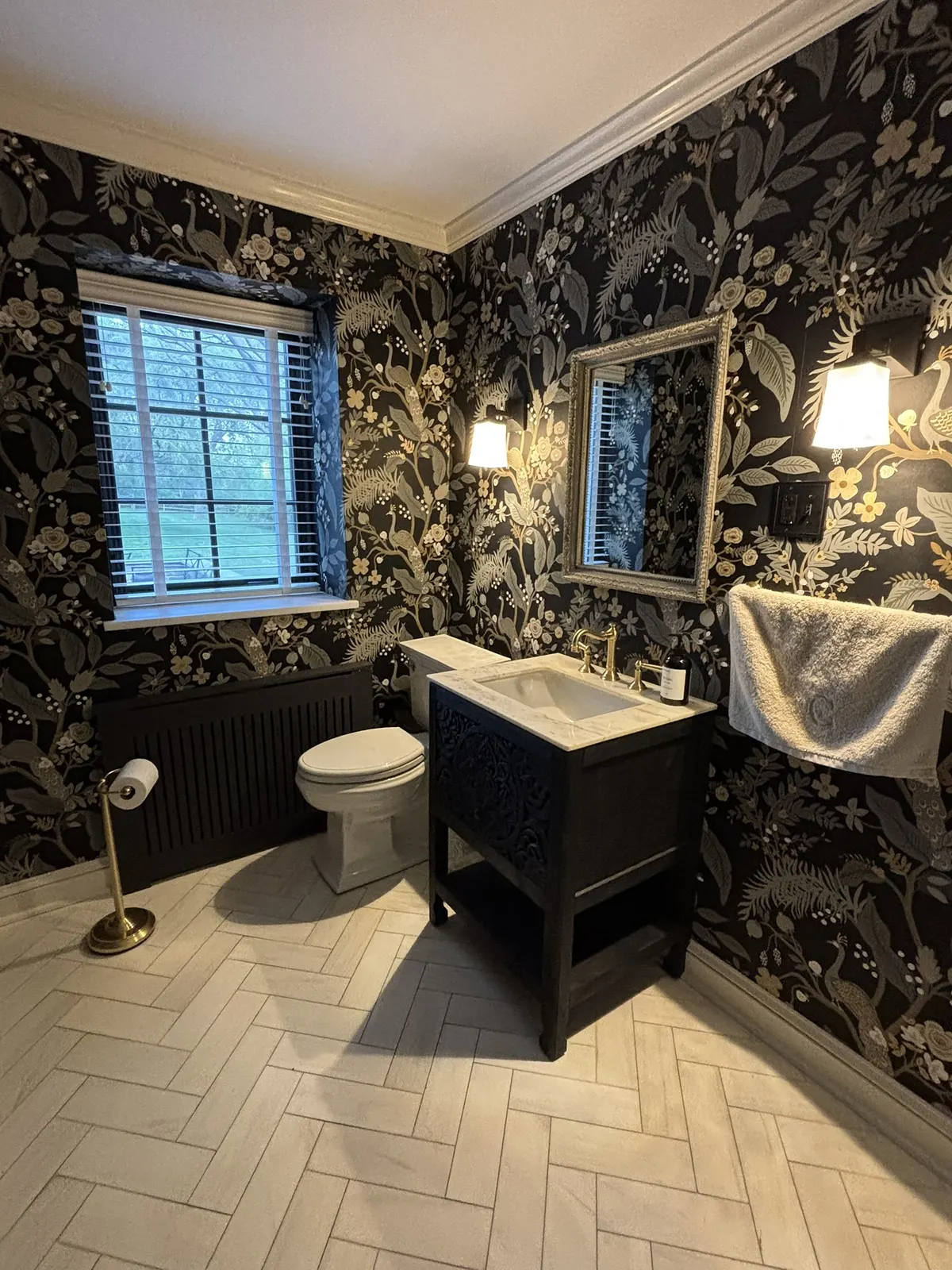Powder room papered in a dark botanical print, with a pale herringbone tile floor, a black furniture-style vanity and a gilt mirror between two sconces.