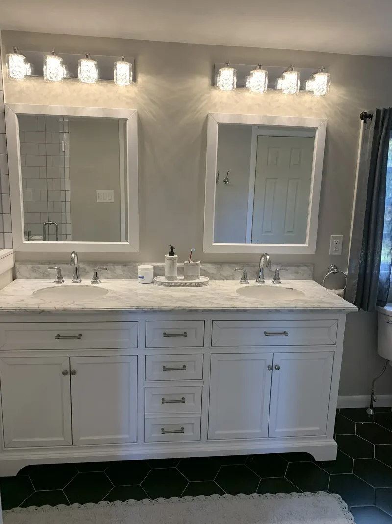 Double white vanity with a marble top, twin mirrors and a row of glass sconces above a black hexagon tile floor.