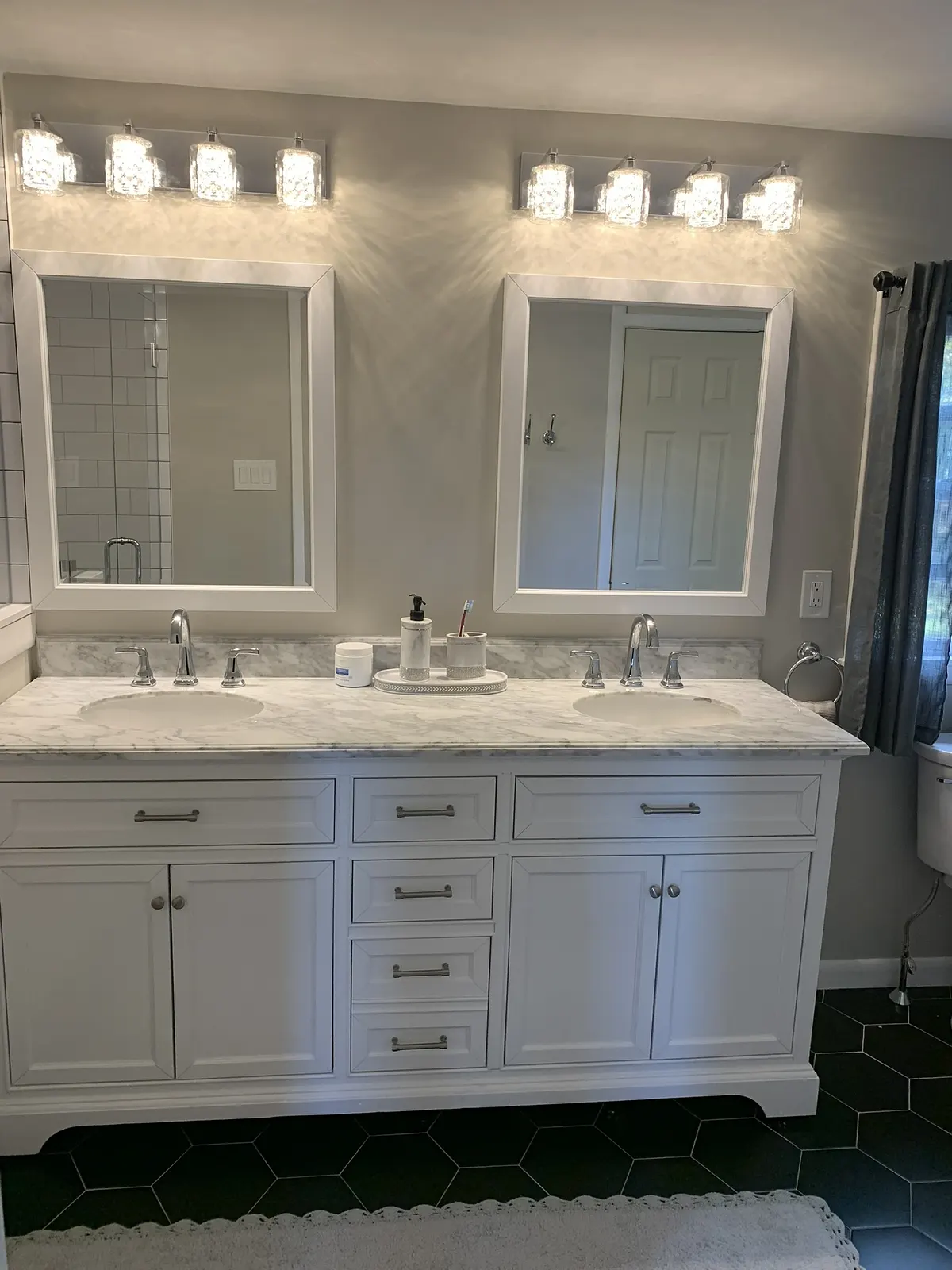 Double white vanity with a marble top, twin mirrors and a row of glass sconces above a black hexagon tile floor.