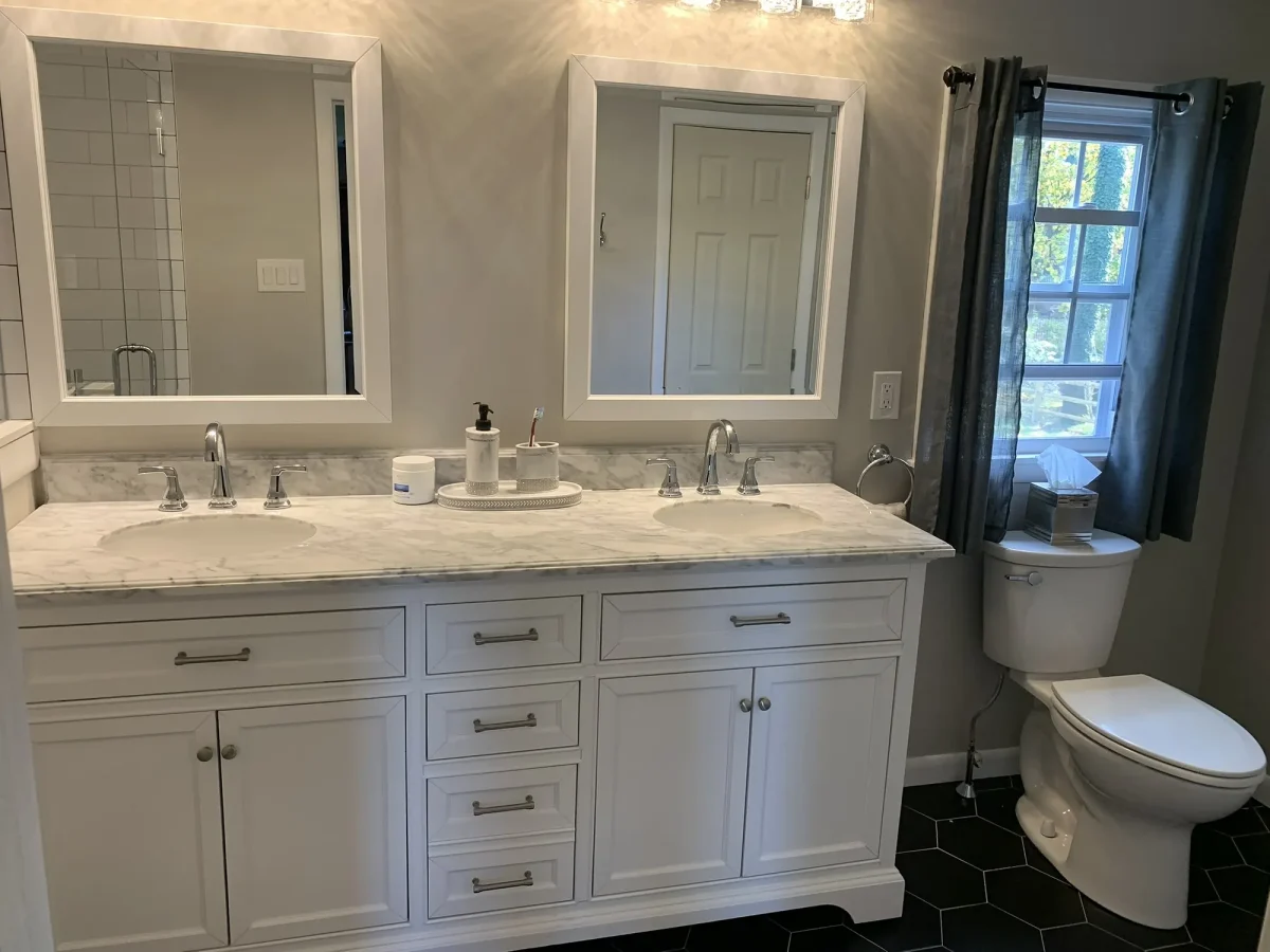 Wide view of the finished bathroom with white cabinetry, marble counter and a window framed in dark curtains.