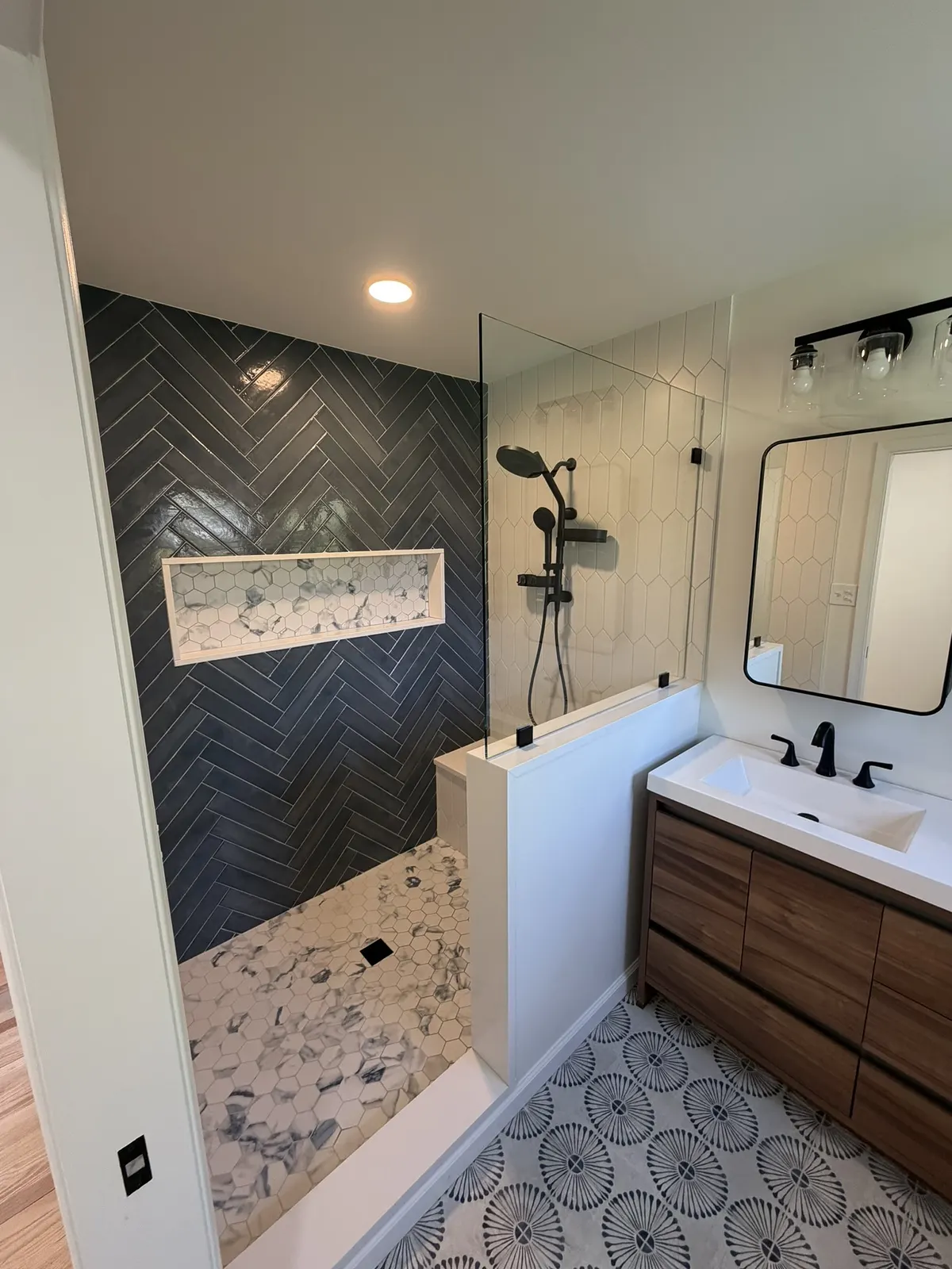 Bathroom with a dark herringbone-tiled shower wall, a walnut floating vanity and a patterned grey-and-white floor.