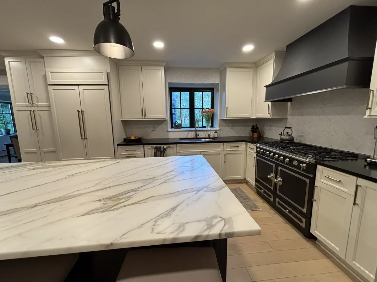 Broad marble-topped kitchen island in the foreground with a black professional range and custom hood on the wall beyond.