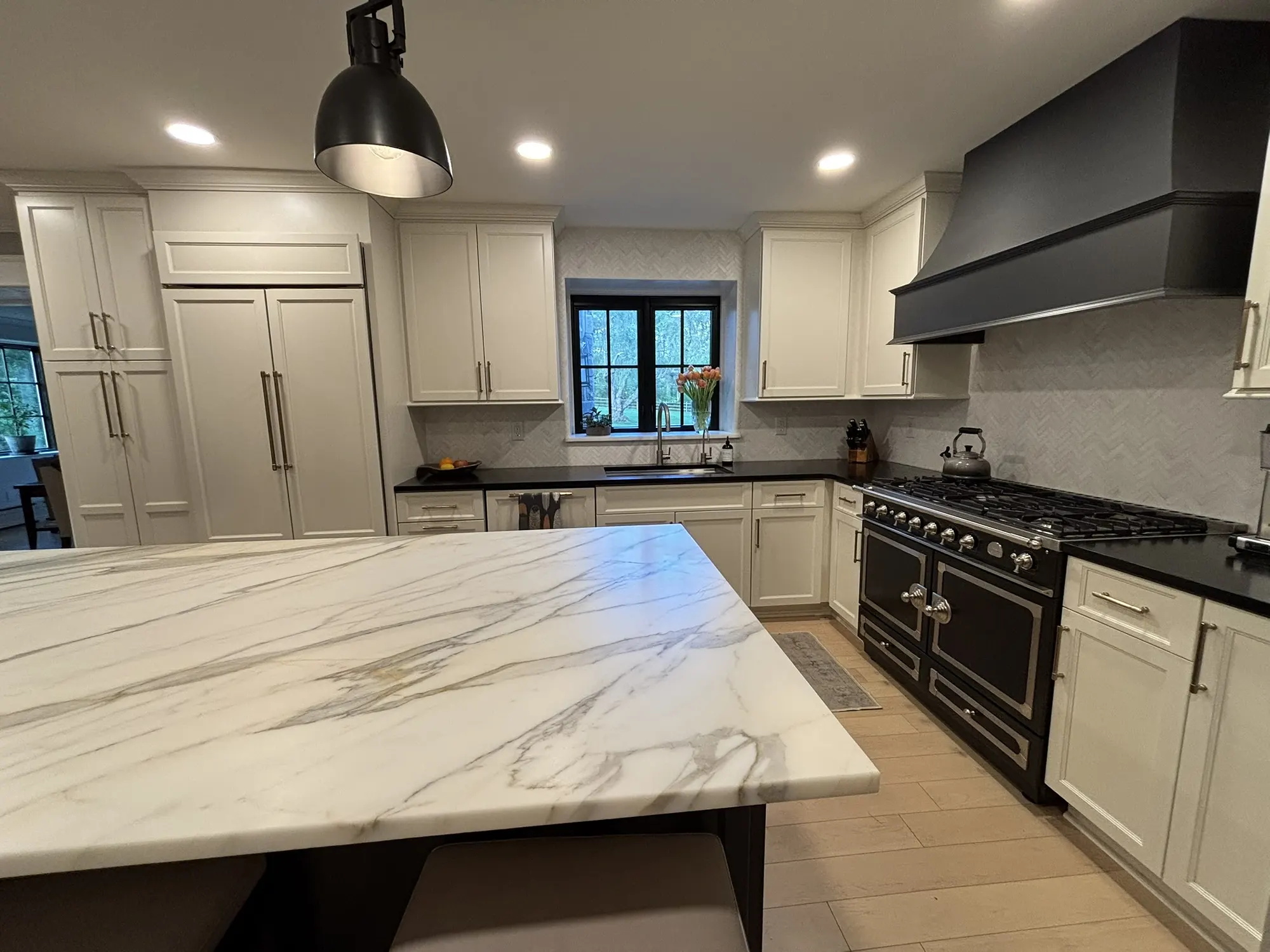Broad marble-topped kitchen island in the foreground with a black professional range and custom hood on the wall beyond.