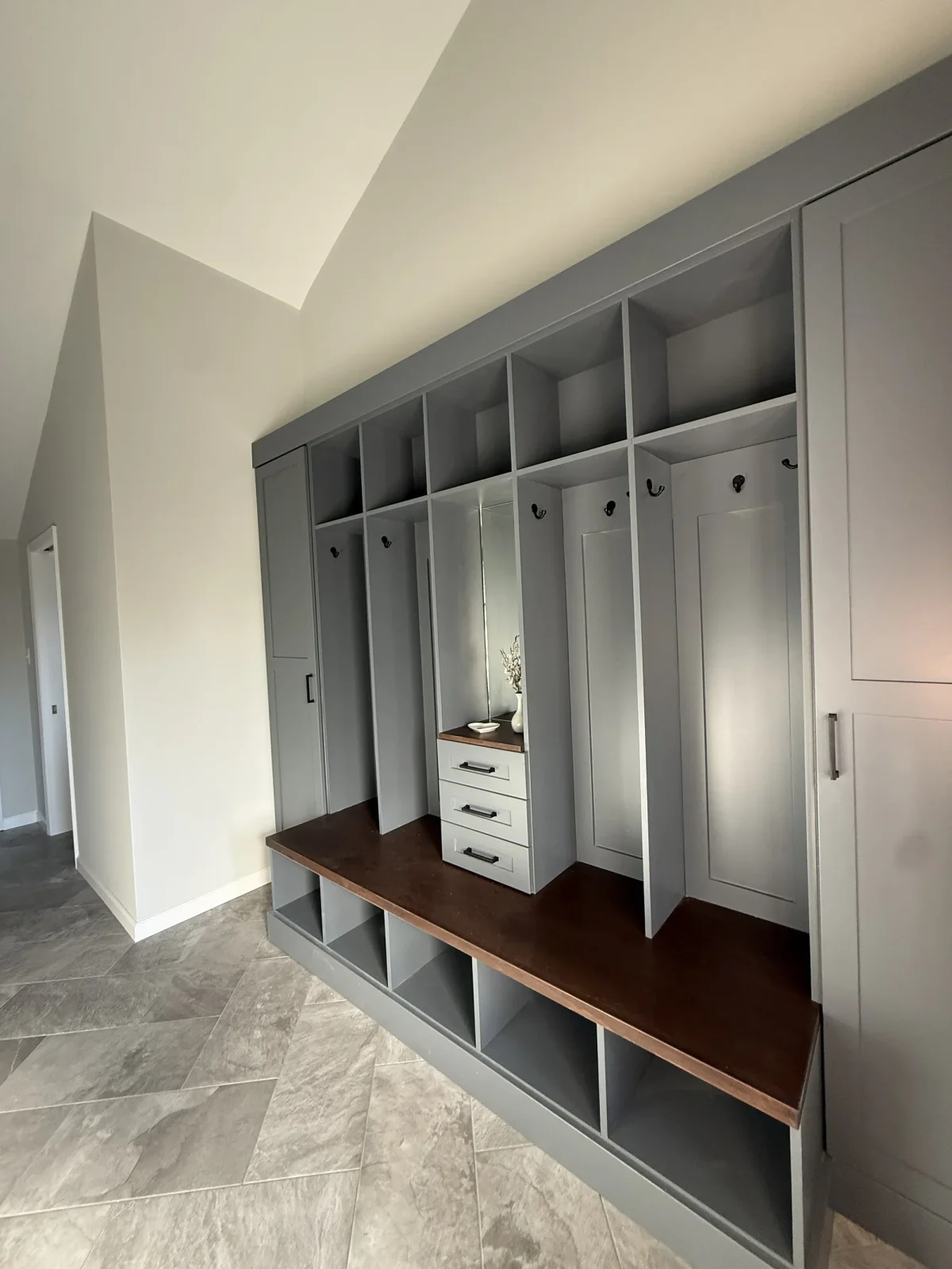 Grey built-in locker bays with a walnut bench top and open cubbies beneath, on large-format tile.