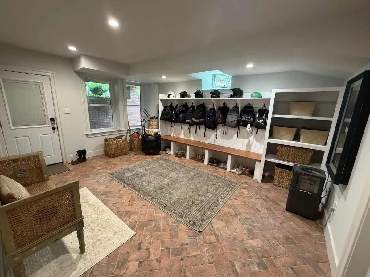 Long run of white built-in mudroom lockers with bench seating and coat hooks, over a herringbone brick floor with a patterned runner.