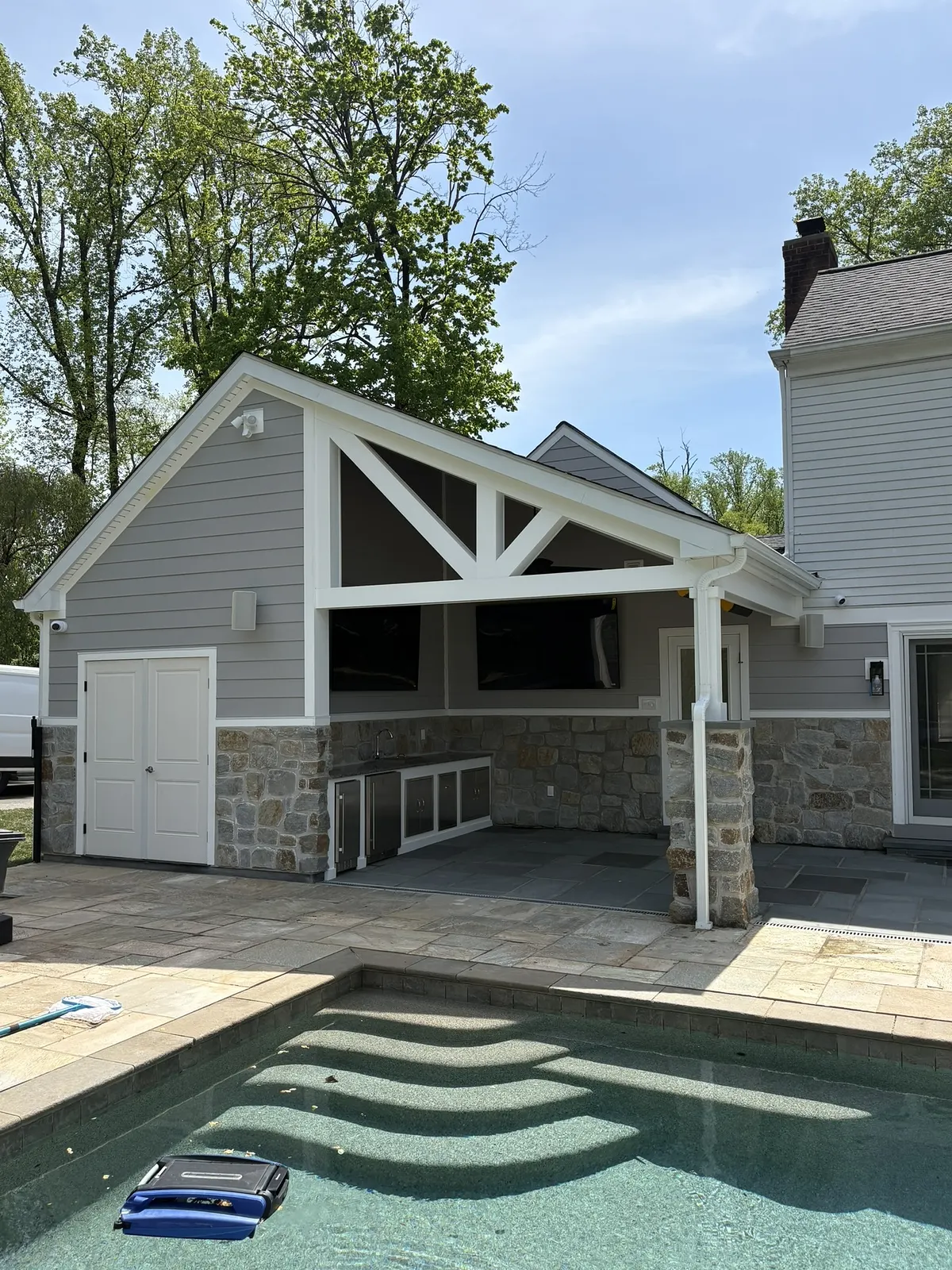 Stone and shingle pavilion with an exposed timber truss standing at the end of a swimming pool, bluestone deck wrapping the water's edge.