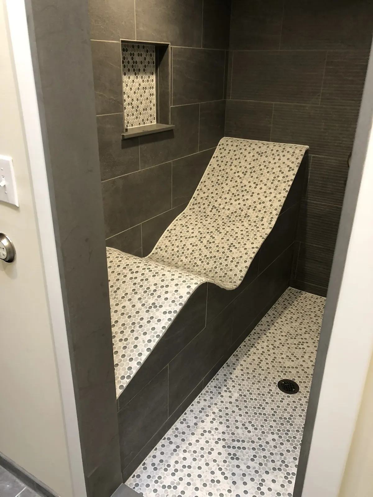 Grey porcelain wet room with a curved tiled chaise longue built into the wall, finished in cream pebble mosaic over a matching pebble floor.
